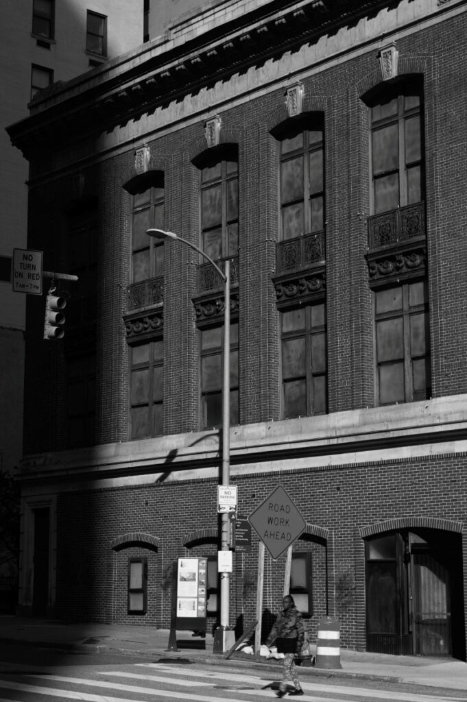 Black-and-white urban street scene with a tall brick building, streetlight, and a 'Road Work Ahead' sign near construction cones on the sidewalk.