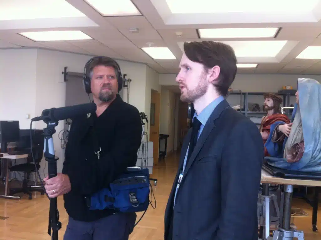 Two men in a recording studio; one wearing headphones speaks into a microphone while another in a suit watches nearby.