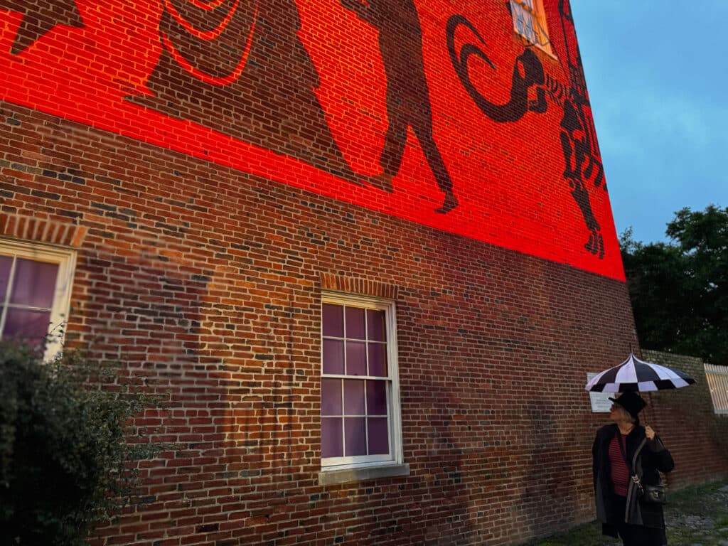 Brick building wall with a large red mural of running silhouettes, blue sky to the right, and a person with a striped umbrella at the bottom right.