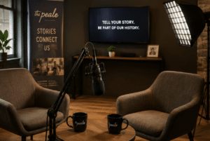 Podcast studio with two gray armchairs around a round table, microphone on a stand, and a screen that says 'Tell your story. Be part of our history.'