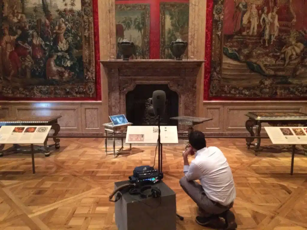 Museum gallery with ornate tapestries and a marble fireplace; a person crouches beside filming equipment.