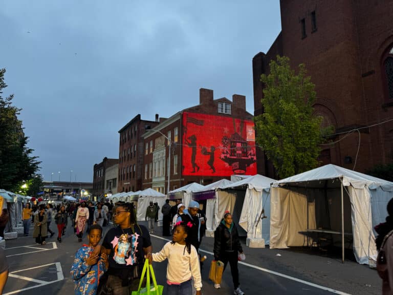 Crowd walking through an outdoor street market with white vendor tents and a red-brick building featuring a large red mural on the right side of the scene.
