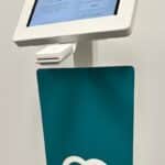 Self-service touchscreen kiosk with a white frame and teal panel bearing a white heart logo.