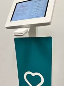 Self-service touchscreen kiosk with a white frame and teal panel bearing a white heart logo.