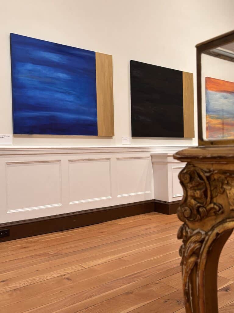 Gallery wall with three large abstract paintings, each with a vertical wooden edge, left-to-right blue, black, and orange-toned works with a white wall background.